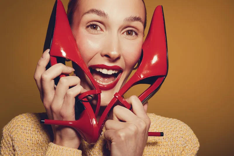 Happy woman with red high heels holds shoes near face smiling widely on yellow background. Concept of fashion, style, footwear, and joyful female emotions in studio lighting.