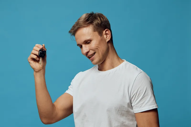 Smiling man with short brown hair, wearing a plain white t-shirt, looking cheerful while holding a black car key in his hand, studio portrait on blue background, lifestyle concept of happiness and suc