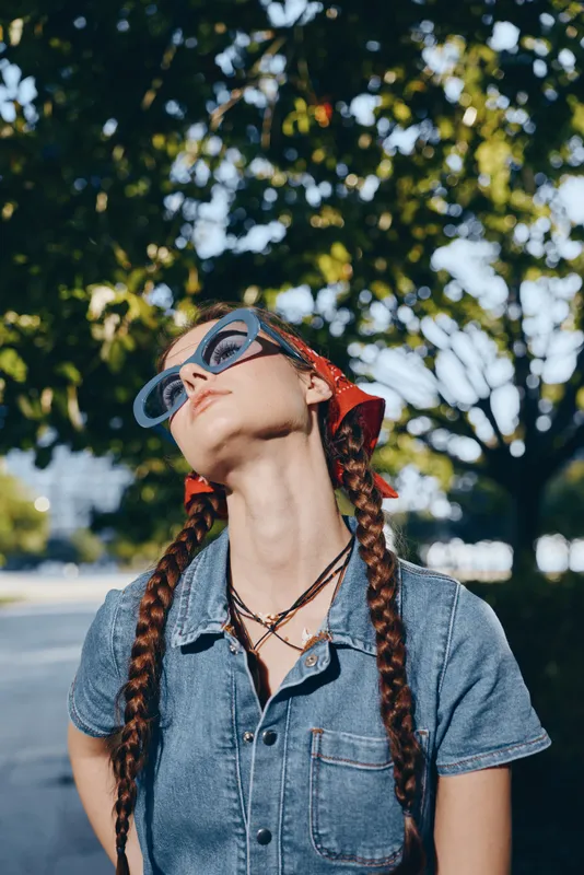 young woman with braids wearing sunglasses and denim shirt outdoors under bright sunlight with green foliage background in warm weather