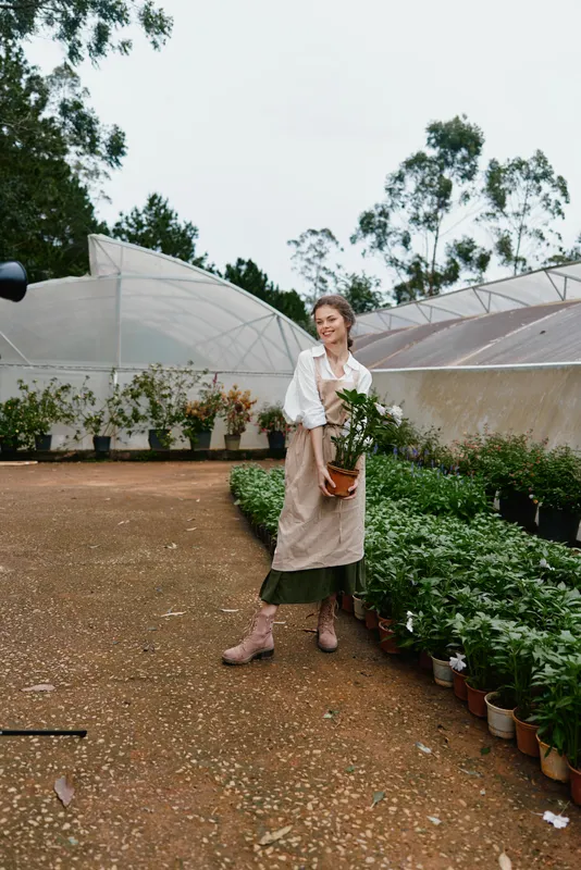 Young woman in a garden, wearing a beige apron, holding a potted plant, surrounded by greenery in a serene outdoor setting