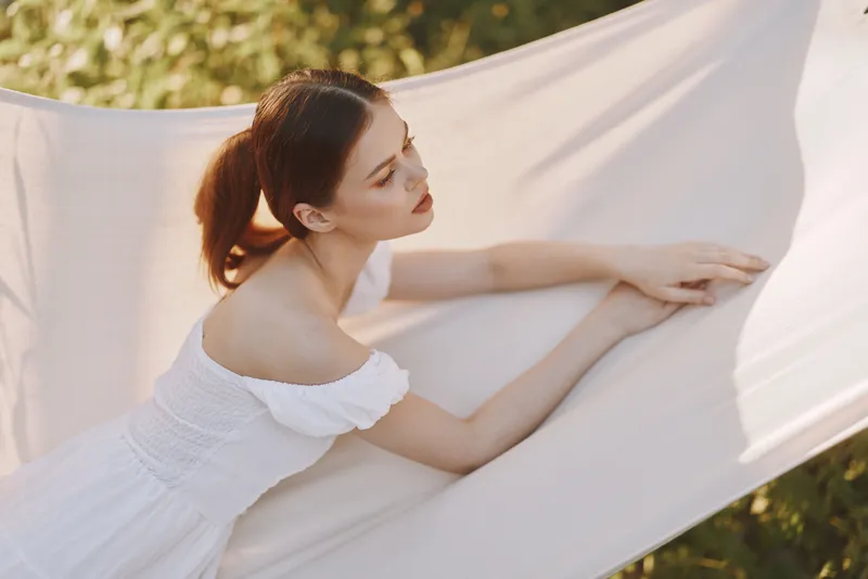 Tranquil young woman in a flowing white dress enjoying serene moments outdoors, enveloped in soft fabric with natural greenery in the background