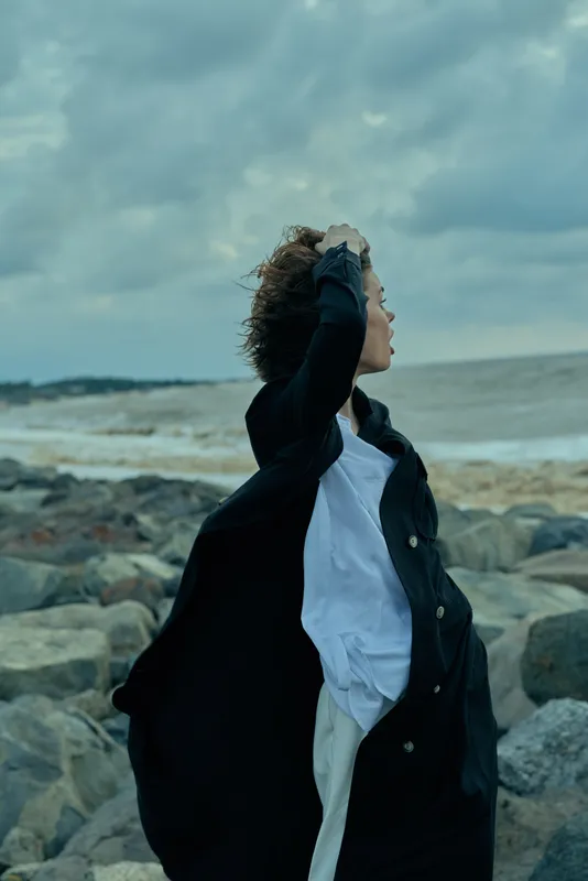 Woman on rocky shoreline wearing a dark coat and white shirt, hair blown by wind as she gazes toward the sky, moody sea breeze and dramatic clouds above