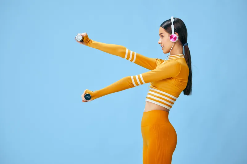 Fitness woman in bright orange athletic wear performing arm exercises with dumbbells, wearing headphones, in a clean studio against a blue backdrop.