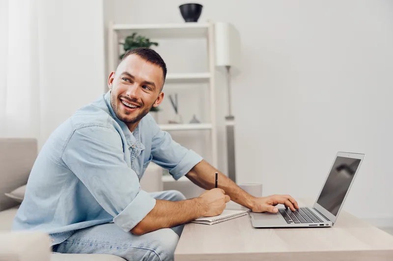 Smiling man working on a laptop in a cozy living room, wearing a light blue shirt, showcasing a modern, relaxed home environment that embodies productivity and comfort.