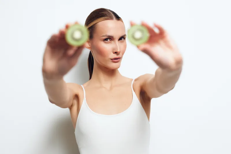 Young woman in white tank top holds two sliced kiwifruits close to camera with focused face isolated on white background, showing fresh healthy lifestyle and natural food concept.