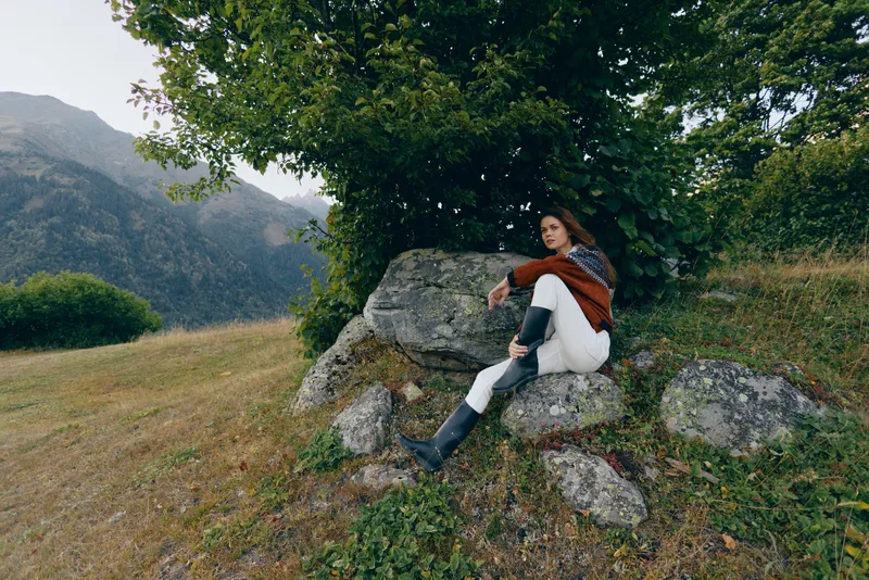 Woman in nature by mountains and meadow, sitting on rocks under a tree for quiet outdoors relaxation; stylish young adult in sweater and boots enjoying peaceful landscape and contemplation.