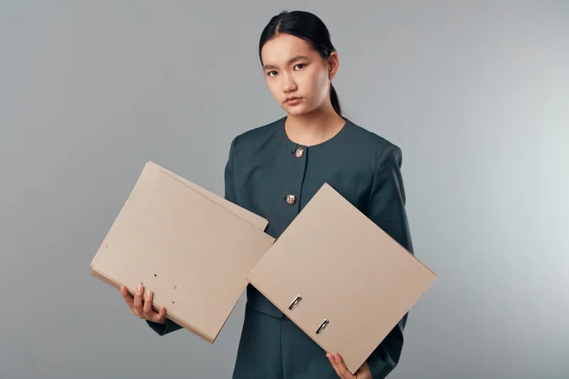 Young woman holding two cardboard boxes with serious expression in studio. Female model with dark hair tied back wearing dark casual outfit against neutral background.