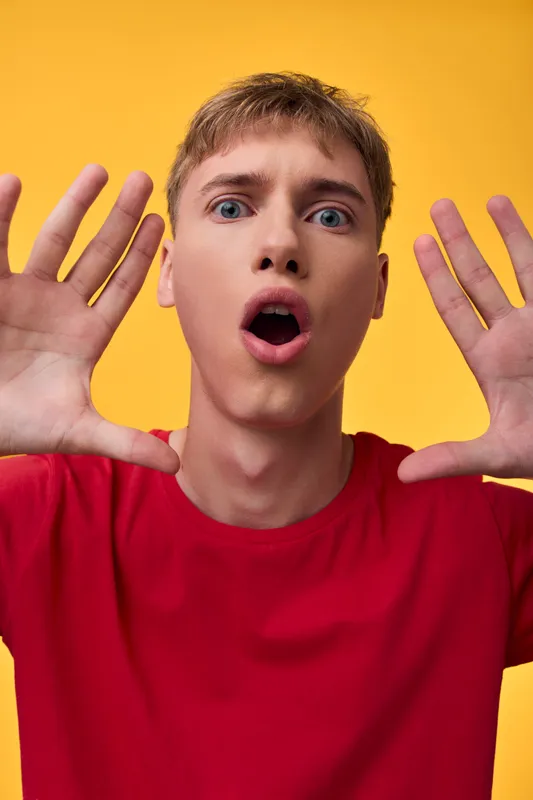 Surprised man in red shirt raises hands toward camera against a bold yellow background, conveying playful astonishment and curiosity, ideal for marketing, ad campaigns, or social media scenes needing