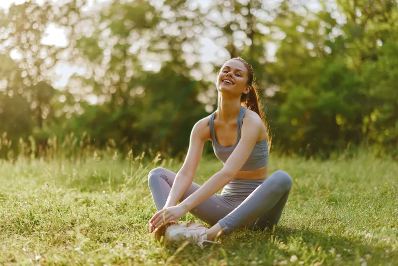 Young woman practicing yoga outdoors in a sunny park, wearing comfortable athletic wear, showcasing a joyful expression amidst greenery and soft sunlight