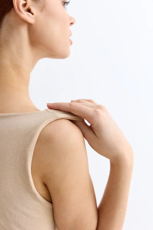 Woman adjusting beige sleeveless top, close-up on shoulder and hand, soft natural skin tones with clean white background for minimalistic style.
