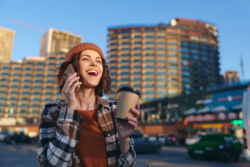 Woman holding coffee cup and phone in city street, smile and candid expression capturing authenticity and candid lifestyle, bathed in golden hour glow for mindful living and emotional storytelling.