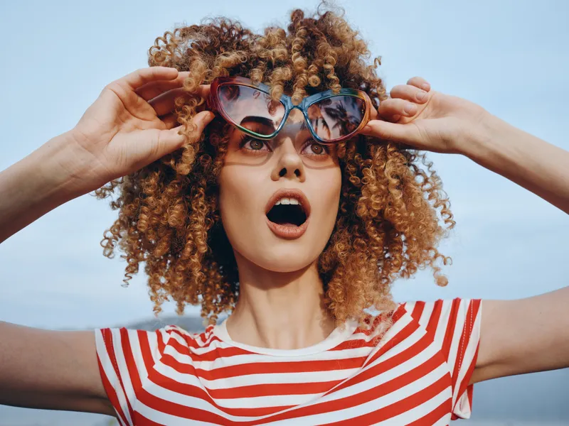 Vibrant portrait of a young person with curly hair wearing a red and white striped shirt and oversized sunglasses, captured with a surprised, playful expression.