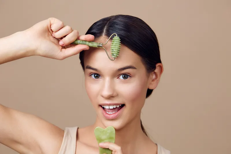 Young woman using a jade roller for skincare, showcasing a joyful expression, with a neutral background and a focus on healthy beauty routines