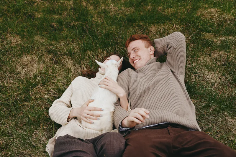 A young couple lies on the grass wearing cozy sweaters while holding and cuddling a small white baby goat in a natural outdoor setting during autumn.