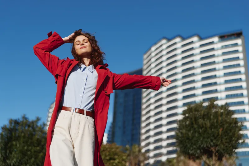 A confident woman in a bright red coat stands outdoors on a sunny day, eyes closed, arms outstretched. Urban architecture in the background highlights bold street fashion.