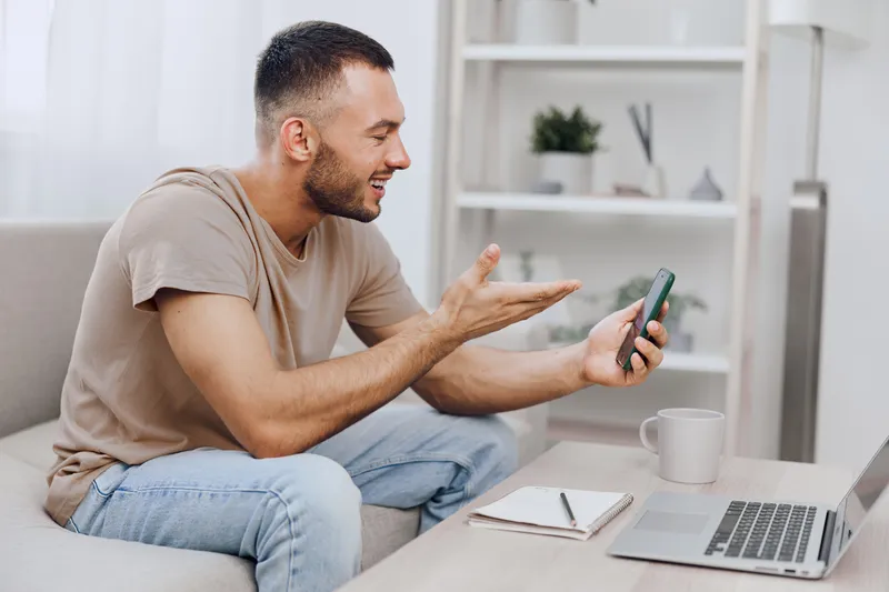 Happy man enjoying a video call while relaxing at home, wearing casual clothes and seated on a cozy sofa. Modern interior design enhances the vibe.