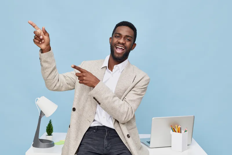 Bright happy cheerful man with dark skin and beard wearing beige blazer and white shirt pointing to side while standing at modern office desk on blue background. People lifestyle concept