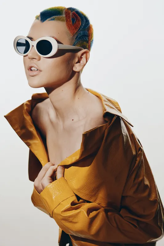 Stylish woman with colorful short hair and white sunglasses wearing a yellow leather jacket, posing confidently against a bright white background in a modern fashion portrait.