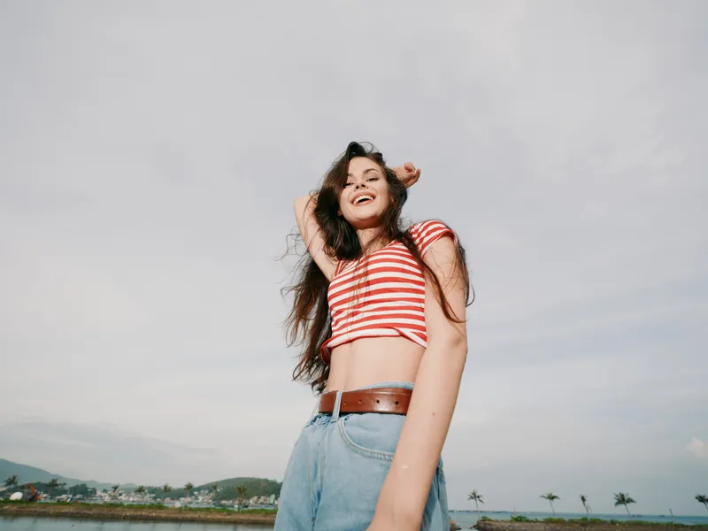 Young woman stands outdoors in a striped red top and denim jeans, smiling confidently at the camera as a soft breeze lifts her hair and sleeves, creating a lively summer moment.