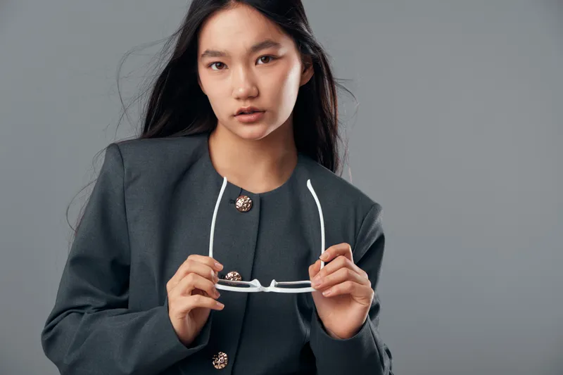 Young woman holding eyeglasses in hand wearing dark gray blazer standing against neutral background with serious expression and straight hair in studio portrait.
