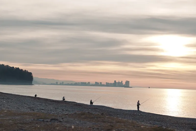 Beach scene at sunset with fishermen along a tranquil shore, calm water and distant city skyline, warm light reflects on waves, creating a peaceful coastal moment.