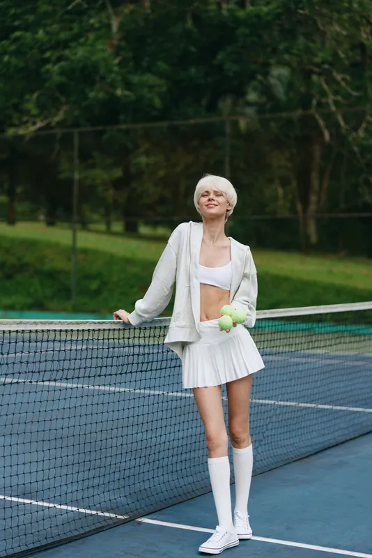 Young female tennis player with short blonde hair in a white sports outfit smiling while holding a tennis ball on an outdoor court surrounded by greenery