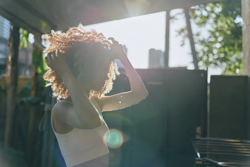silhouette of woman with curly hair adjusting her hairstyle outdoors in sunlight, casual outfit, natural light, urban background, bright and fresh atmosphere