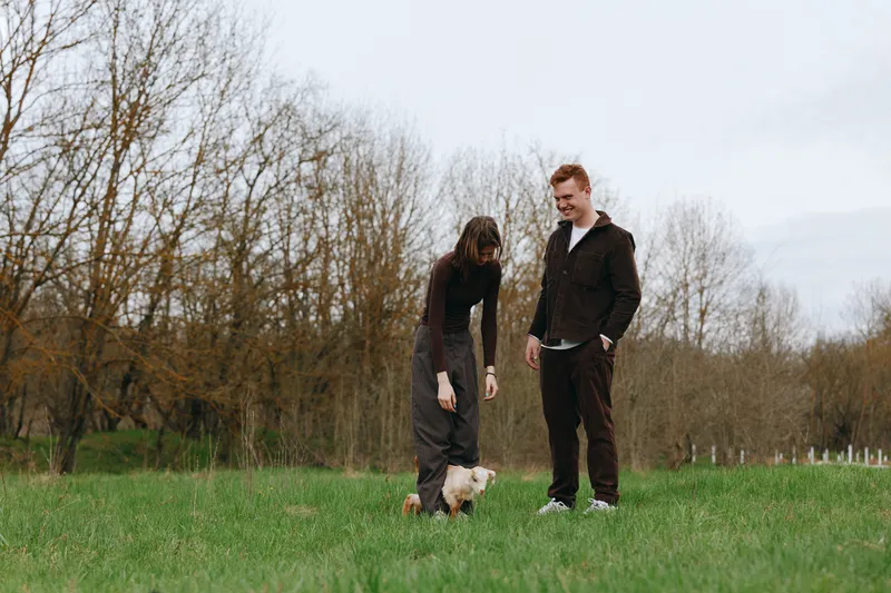 A young woman and man enjoy a casual moment outdoors while playing with a small white goat in a green grassy field under an overcast sky during early spring.