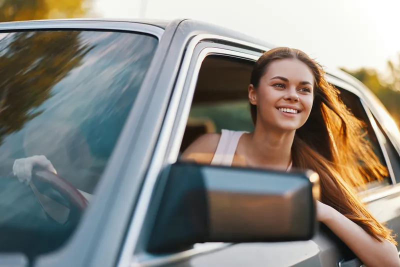 Smiling young woman enjoying a road trip with long hair flowing in the wind from the car window in a sunny landscape, evoking a sense of freedom and adventure.