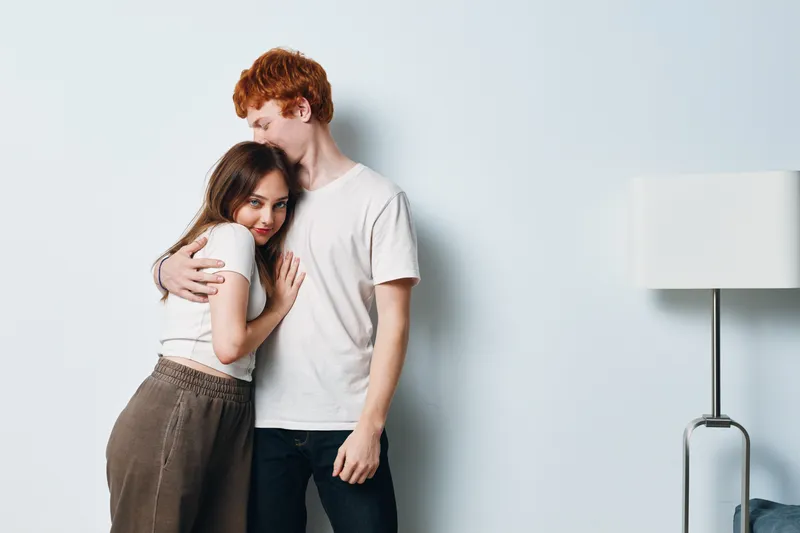 Couple embracing and smiling in casual clothes at home, standing near wall and modern lamp, young man and woman in comfortable lifestyle and relationship moment indoors.