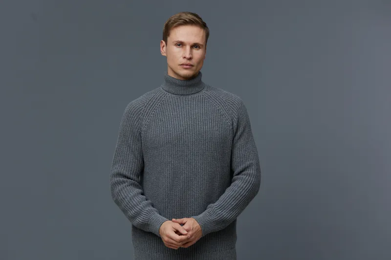 Male model in a cozy gray turtleneck sweater looking serious and contemplative, standing against a plain dark gray background. Casual fashion concept, studio portrait, fashion styling, autumn or winte
