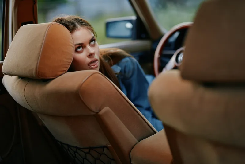 Young woman with casual clothing, looking playfully over the car seat, with a relaxed expression, outdoors, showcasing a summer vibe in warm colors.