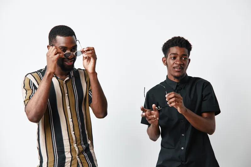 Two young black men with dark skin posing with sunglasses on white background. Casual style, confident expressions, and modern fashion with striped and black shirts.