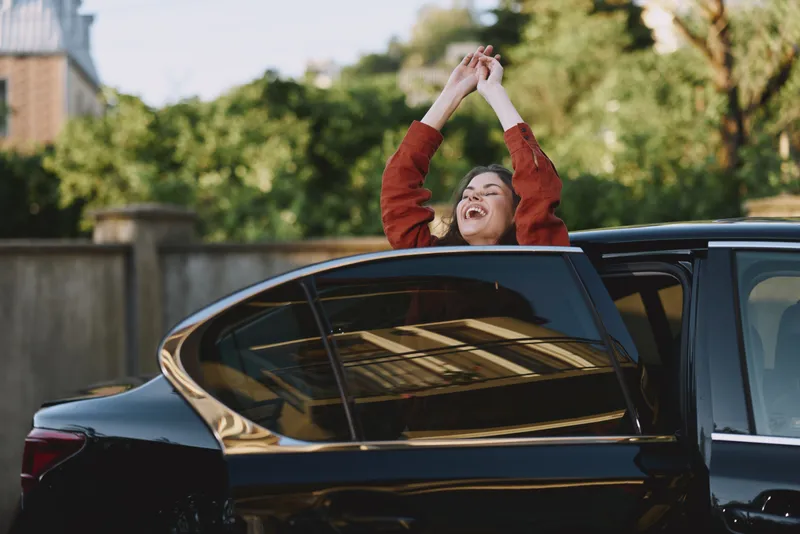 A joyful woman exits a sleek car with arms raised, face lit with triumph. An outdoors candid moment capturing celebration, happiness, and a vibrant lifestyle scene