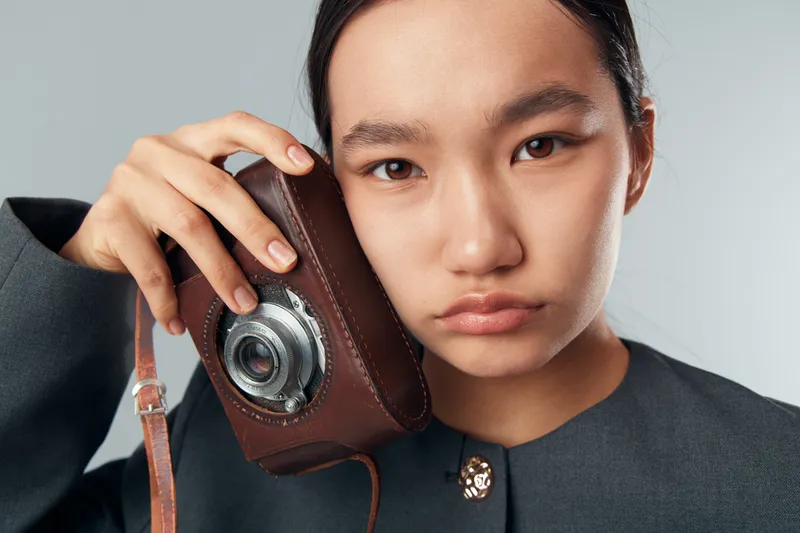 Young woman holding vintage camera close to face looking at viewer with serious expression. Portrait of female photographer with retro equipment and brown leather case in studio setting.