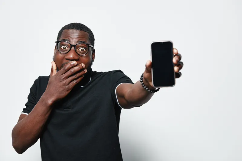 African American man with dark skin and glasses showing smartphone with blank screen and surprised expression on face in studio with white background.