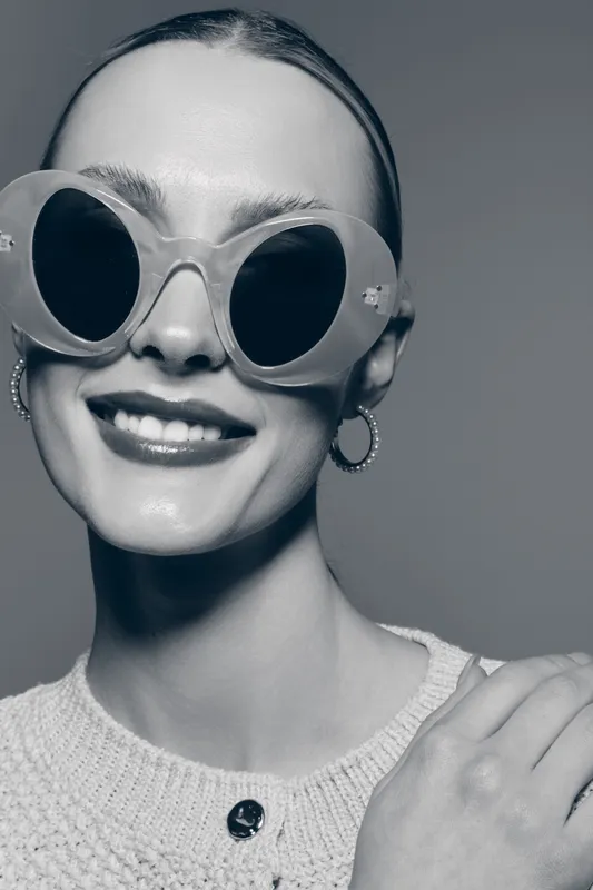 Smiling Caucasian woman with fair skin wearing large round sunglasses and hoop earrings in black and white close-up.