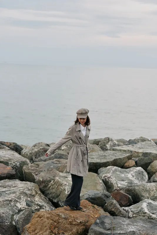 Woman in warm coat and hat stands on rocky shore by calm sea under cloudy sky. Winter fashion and outdoor lifestyle concept with natural coastal background and relaxed mood.