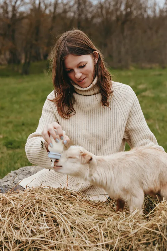 A woman feeds a young goat outdoors, seated on straw in a peaceful field. A candid lifestyle moment showcasing care, gentleness, and daily connection with animals.