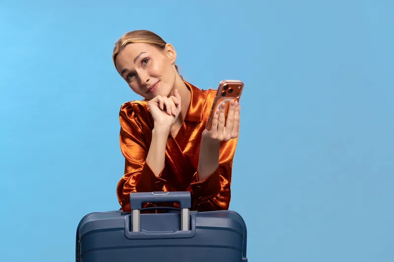 Thoughtful woman in orange satin blouse holding smartphone leans on blue suitcase against blue background. Concept for travel planning, vacation ideas, and leisure time activities.