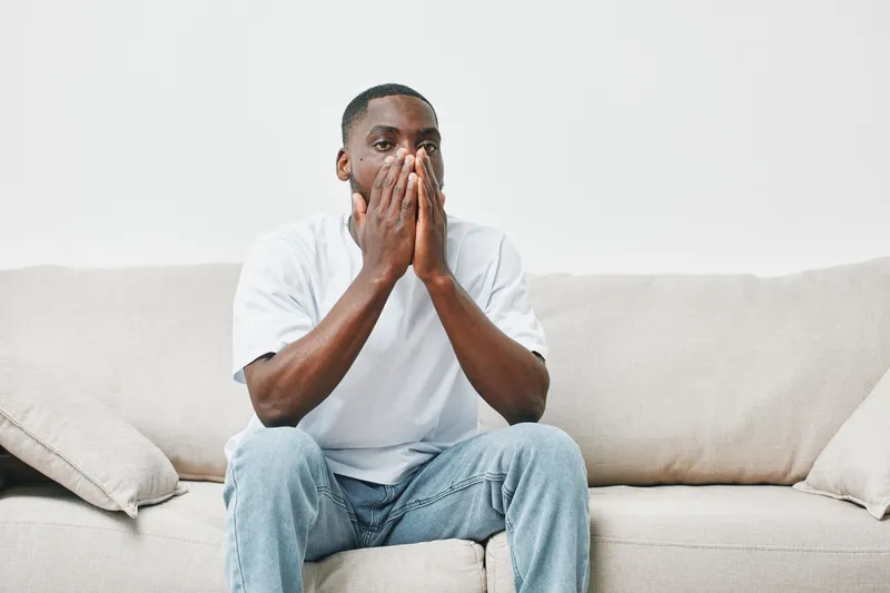 A pensive young African American man in a white t shirt sits on a beige sofa, reflecting on life s challenges with a thoughtful expression