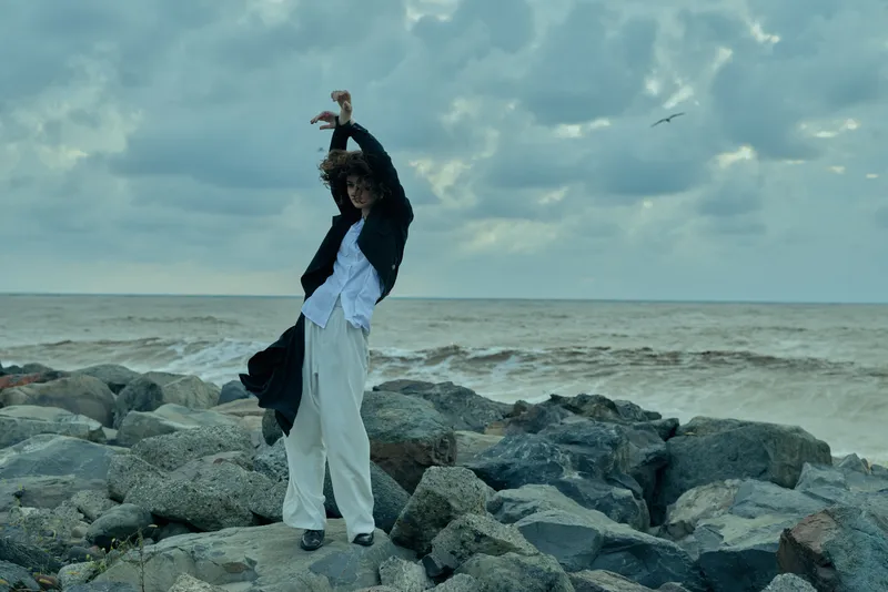 A woman dances on a rocky shoreline, flowing white garment in the wind, with sea and cloudy sky behind, capturing movement, freedom, and a dramatic coastal moment