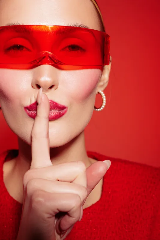 Close-up of woman wearing red glasses and red sweater making silence gesture with finger on lips. Concept of secrecy, quiet, and mysterious mood on red background.