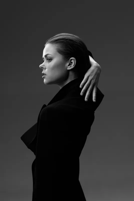 Black and white portrait of a contemplative woman with braided hair in a dark coat, posing elegantly against a minimalist background.