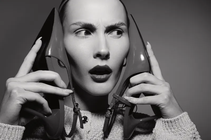 Surprised white woman with pale skin holds red high heels near her ears, looking sideways with open mouth. Black and white studio portrait with emotional facial expression concept.