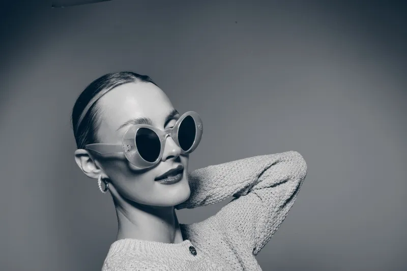 Stylish white woman with pale skin wearing round sunglasses and sweater poses confidently in monochrome studio lighting.