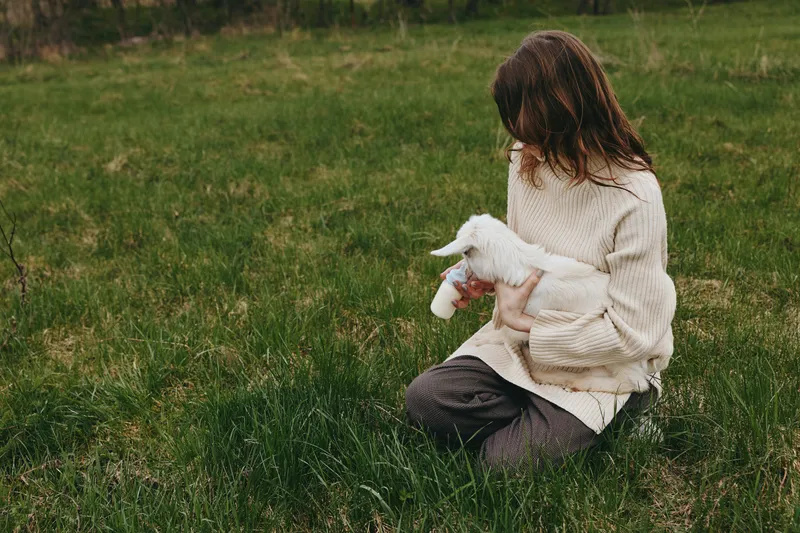 A woman in a cozy sweater sits on grass outdoors holding a young goat kid, sharing a tender moment. Natural light and a calm rural setting emphasize simple lifestyle and gentle bonding with animals.