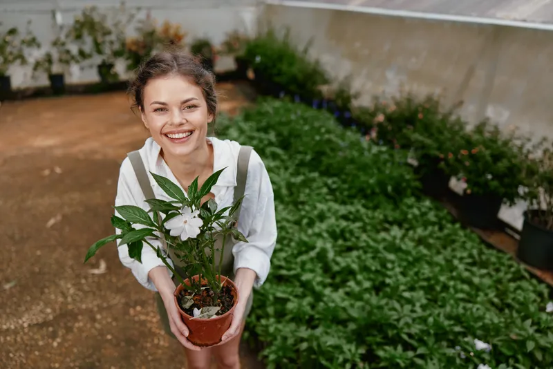 Happy young woman with a plant in a greenhouse, showcasing a love for gardening and nature, surrounded by greenery and flowers