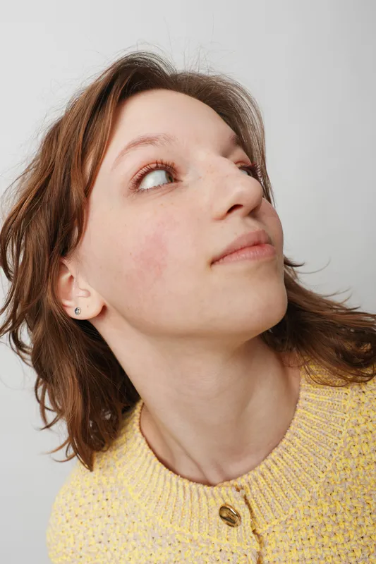 Young woman with short brown hair looks upward with thoughtful expression. She wears a yellow knitted sweater and small earrings against a plain white background. Concept of contemplation.