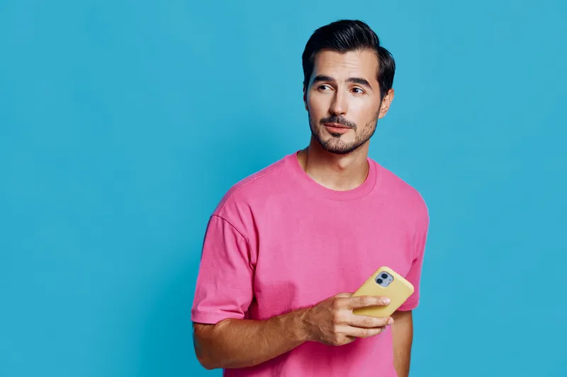 Full body thoughtful young man wearing a pink t-shirt, looking away while holding a smartphone against a bright blue background in a studio setting. People lifestyle concept